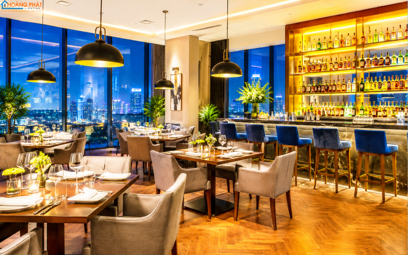 Ảnh nhà hàng khách sạn buổi tối với đèn rọi bàn ăn và quầy bar hắt sáng kệ rượu