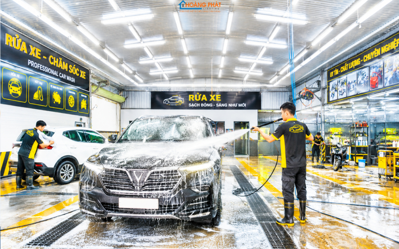 Tiệm rửa xe sáng trưng với dàn máng đèn LED chống ẩm trên trần, nhân viên đang xịt rửa ô tô